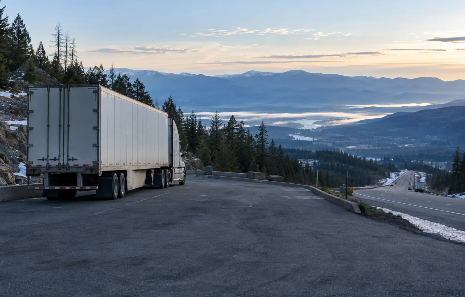 An unbranded tractor-trailer waits safely in a paved mountain turnout above a descending highway at cool-blue dawn