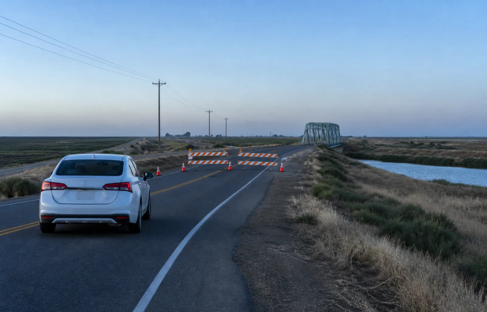 A car stopped before orange barricades on a two-lane Delta highway leading to a small steel bridge